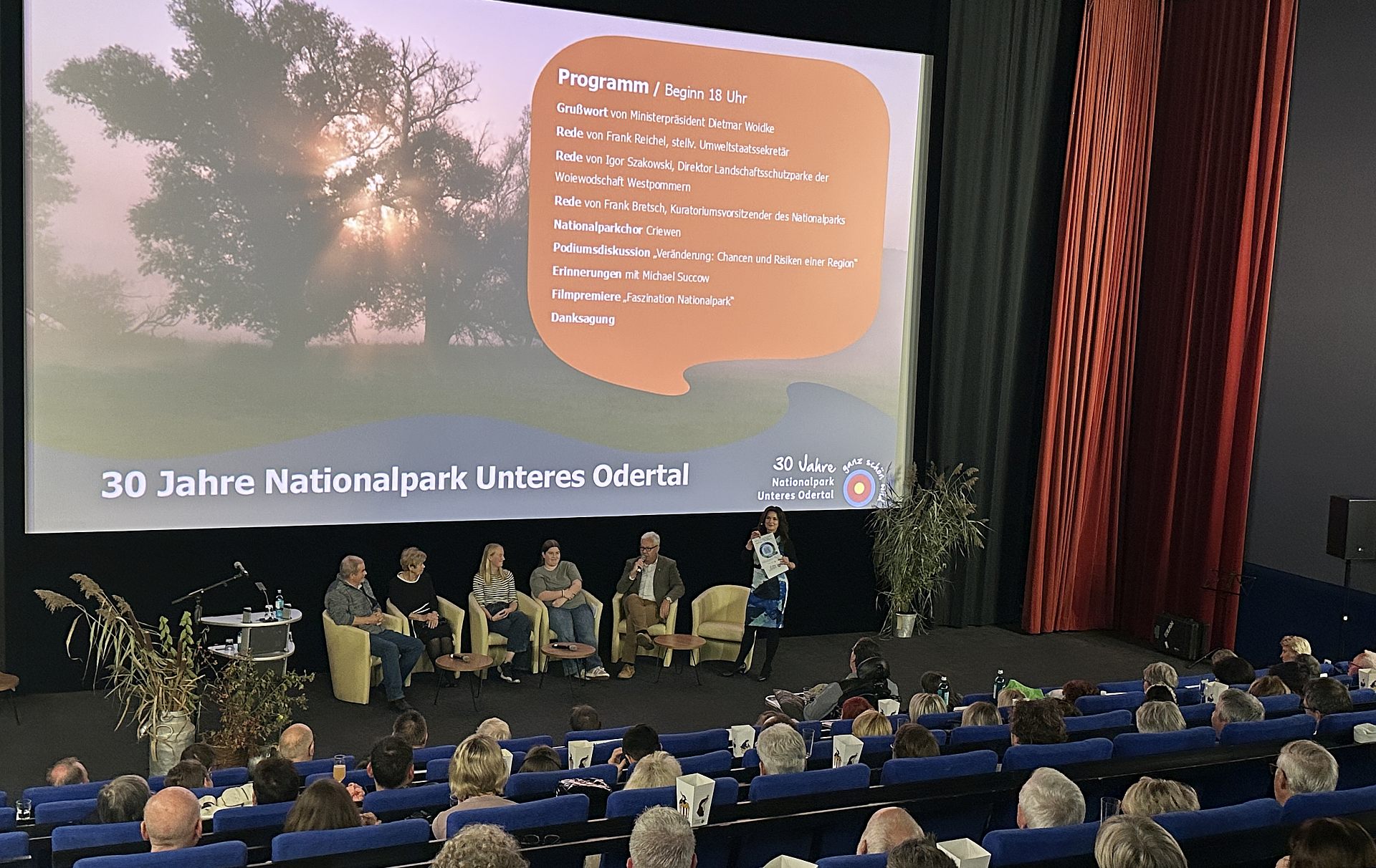 Foto: Kinosaal mit Publikum und einer Podiumsdiskussion vor der Leinweind