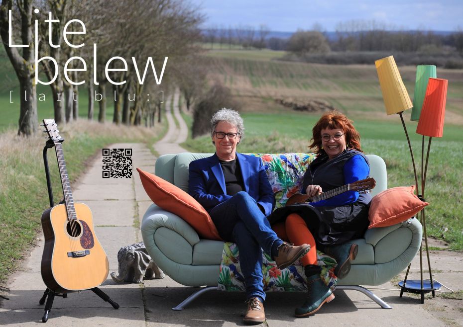 Eins Frau und ein Mann sitzen auf einem Sofa. Das Sofa, die Stehlampe, der Dekoelefant und die Akustikgitarre befinden sich auf einem Plattenweg mitten in der Landschaft. 