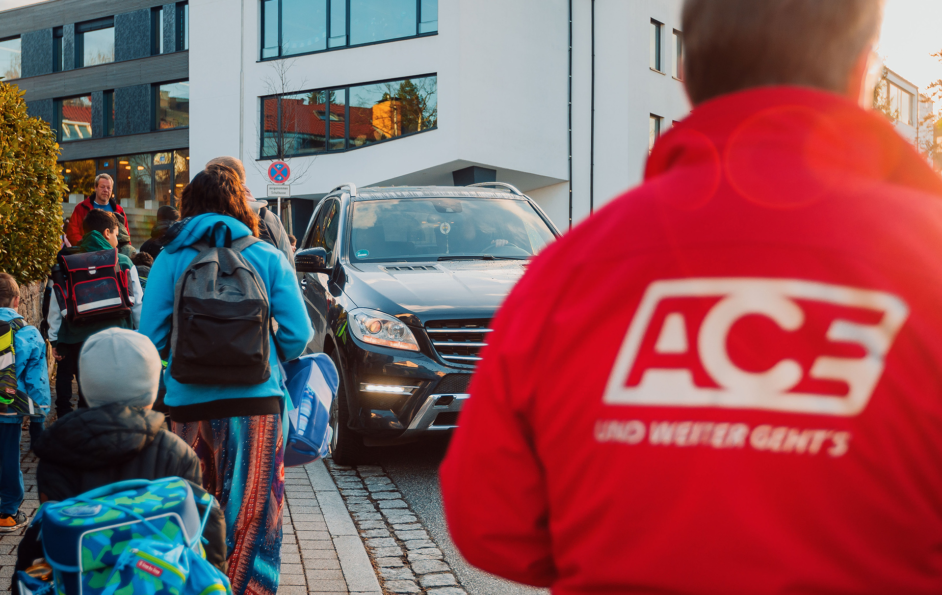 Foto: Schulkinder laufen an einem Auto vorbei, ein Man in roter Jacke beobachtet das