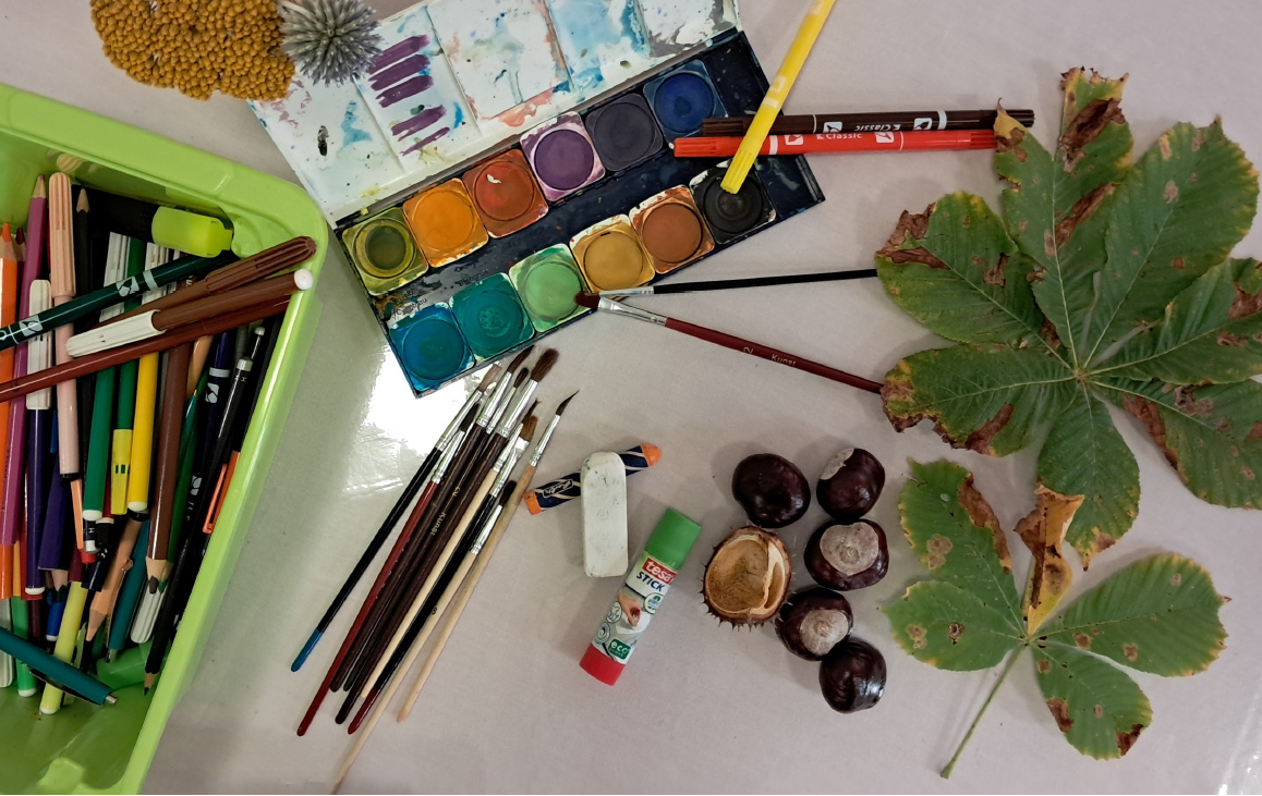 Foto: auf dem Tisch liegen Stift, Pinsel, Tuschkasten, Kleber, Kastaniern und Blätter