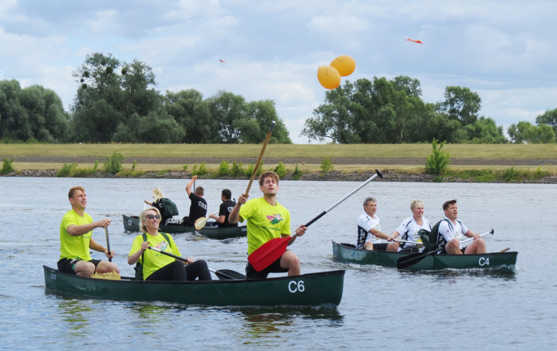 Foto: 3 Boote im Wettstreit, im vorderen Boot versucht ein Sportler stehend einen orangenen Luftballon über sich zu zerstechen.