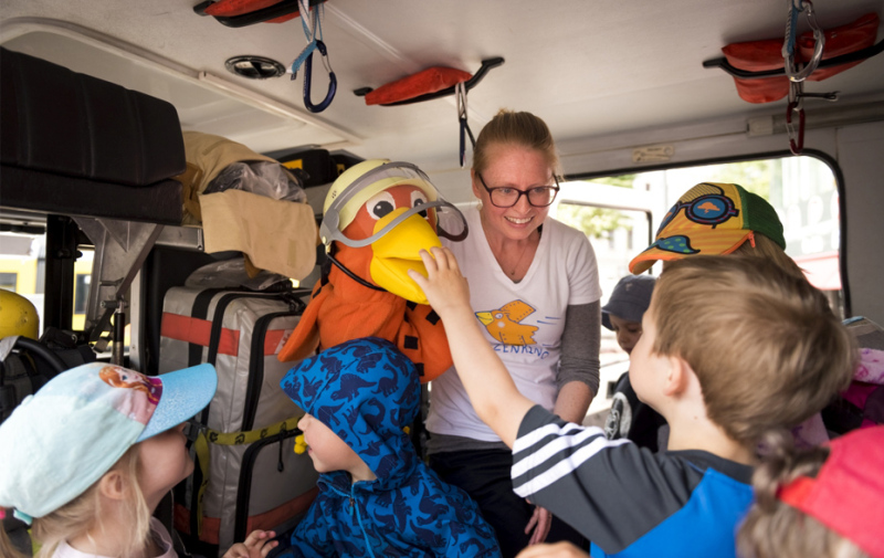 Foto: Frau mit Spatzenpuppe, umringt von Kindern in einer Feuerwehr