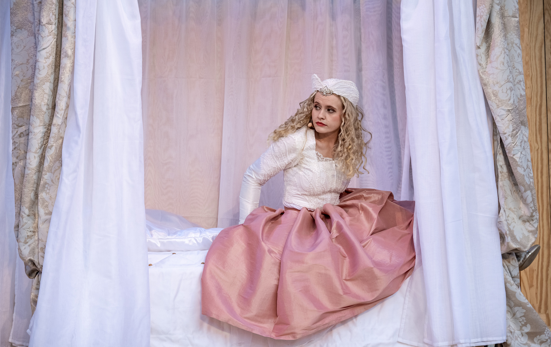 Foto: Schauspielerin in rosa Kostüm sitzt auf einem Bett