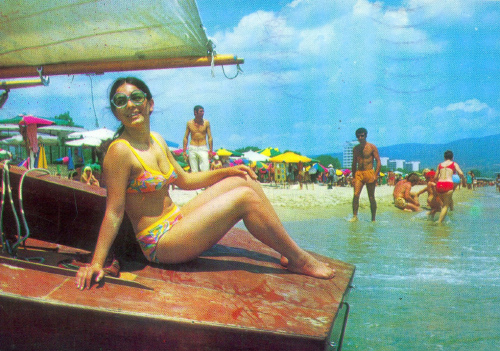 Die Postkarte zeigt eine Frau, auf einem Boot sitzend, am gut gefüllten Badestrand. Die Postkarte zeigt eine Frau, auf einem Boot sitzend, am gut gefüllten Badestrand.